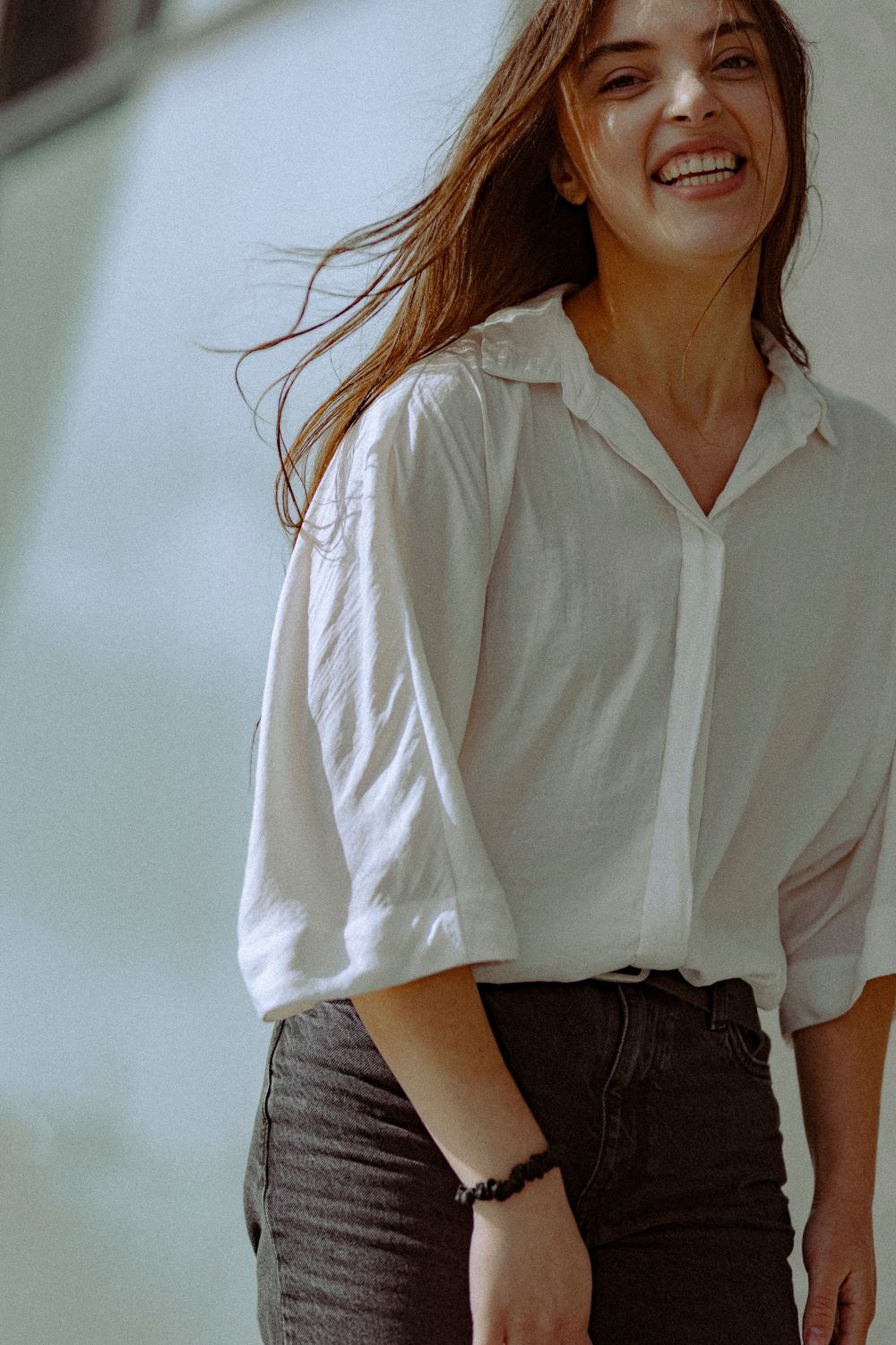 A person in a white button-down shirt and dark jeans smiles with windswept hair against a light background.