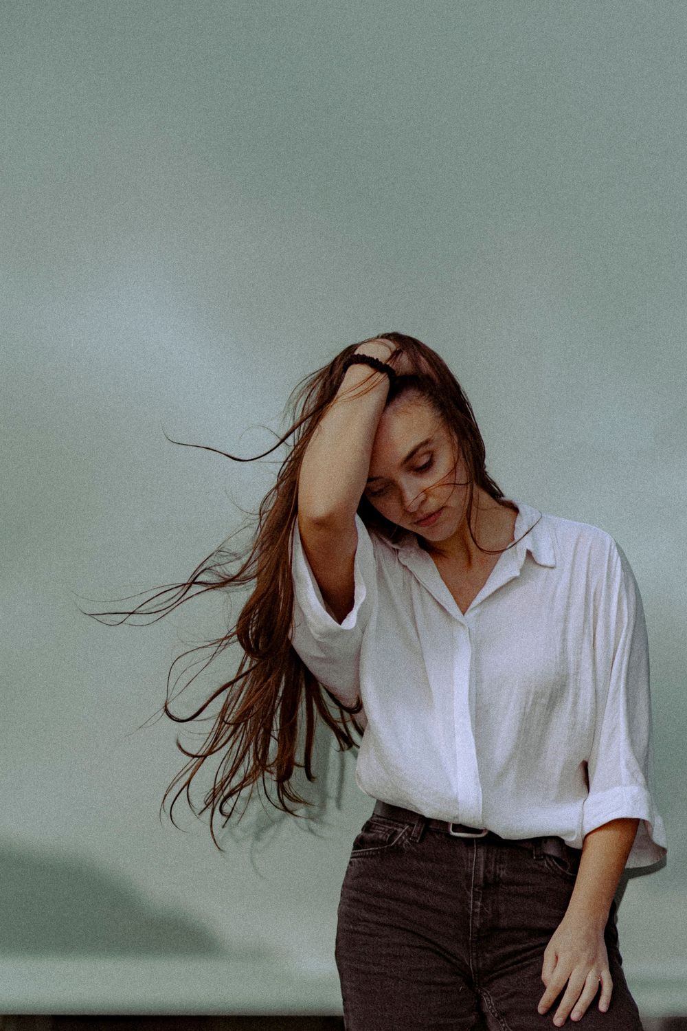 A person with flowing hair poses in a white button-down shirt and dark jeans against a muted green background.