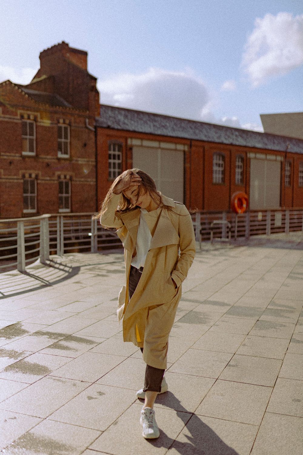 Figure in a beige trench coat walks across a concrete terrace near historic brick buildings.