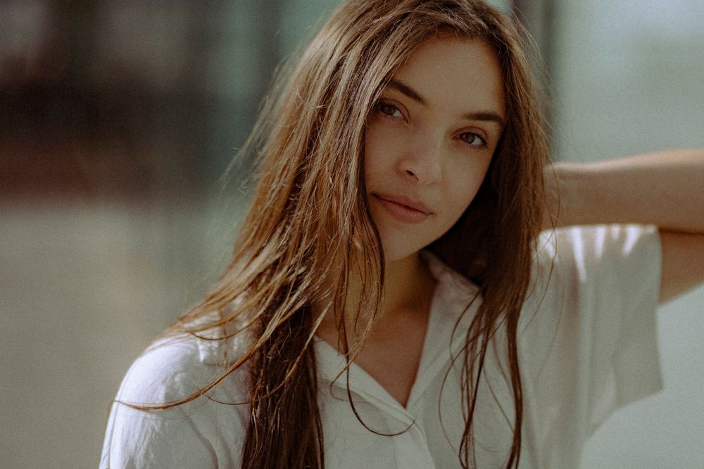 Portrait of a young woman with long dark hair wearing a white top in soft natural lighting.