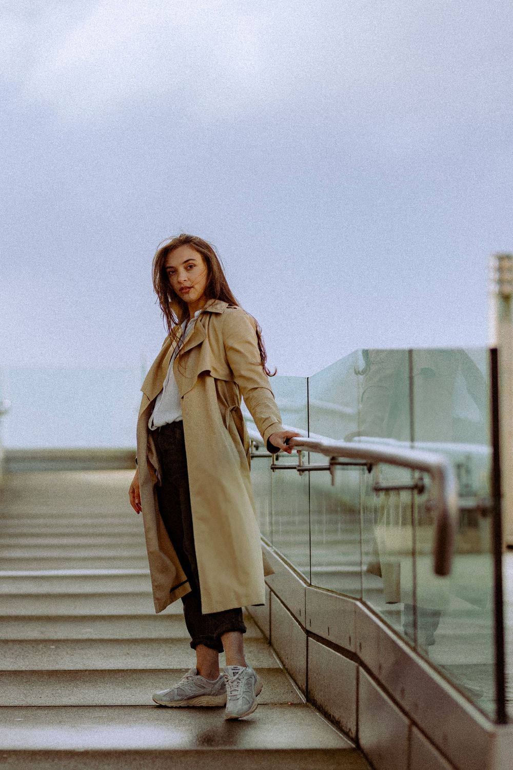 Person in beige trench coat and sneakers poses on concrete steps in moody urban setting.
