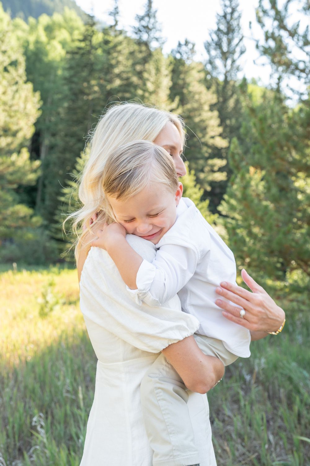 A mother with blonde hair holds her young child in a warm embrace outdoors among green trees and meadow.