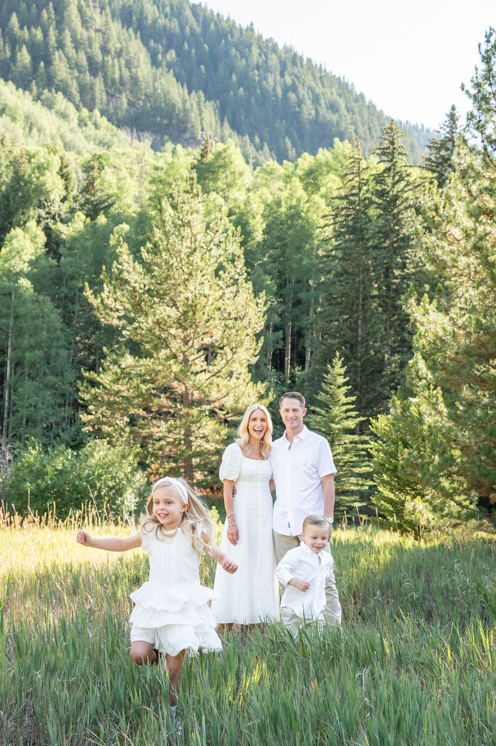 Family in white outfits posing in a green mountain meadow with tall pine trees, children playing in the foreground.