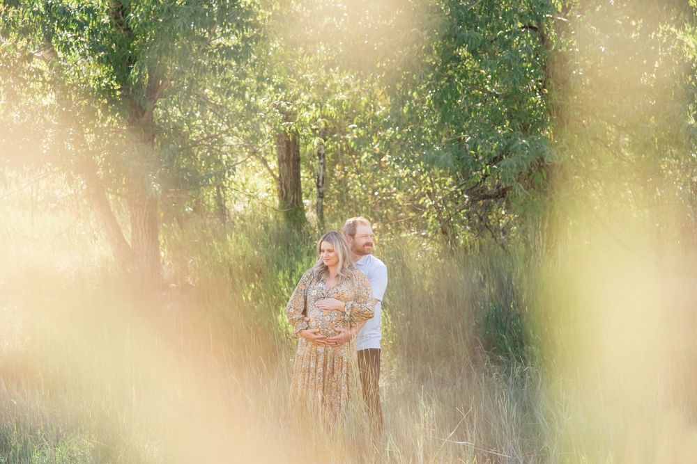 Whimsical photo of couple both holding woman's pregnant belly while in a field with golden grasses and trees behind them in Vail Colorado