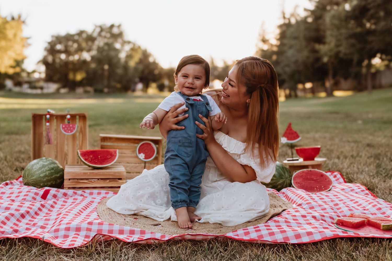 A picnic scene on a checkered blanket with watermelon and summer refreshments at sunset in a grassy park.