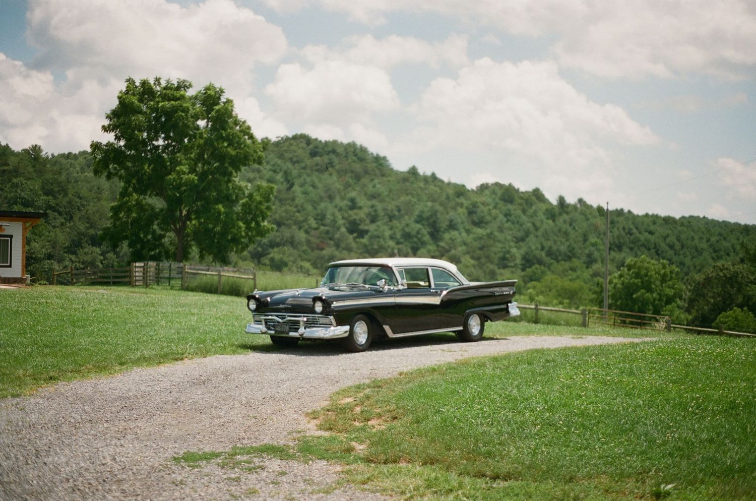 A black 1950s Ford automobile parked on a gravel driveway overlooking rolling hills.