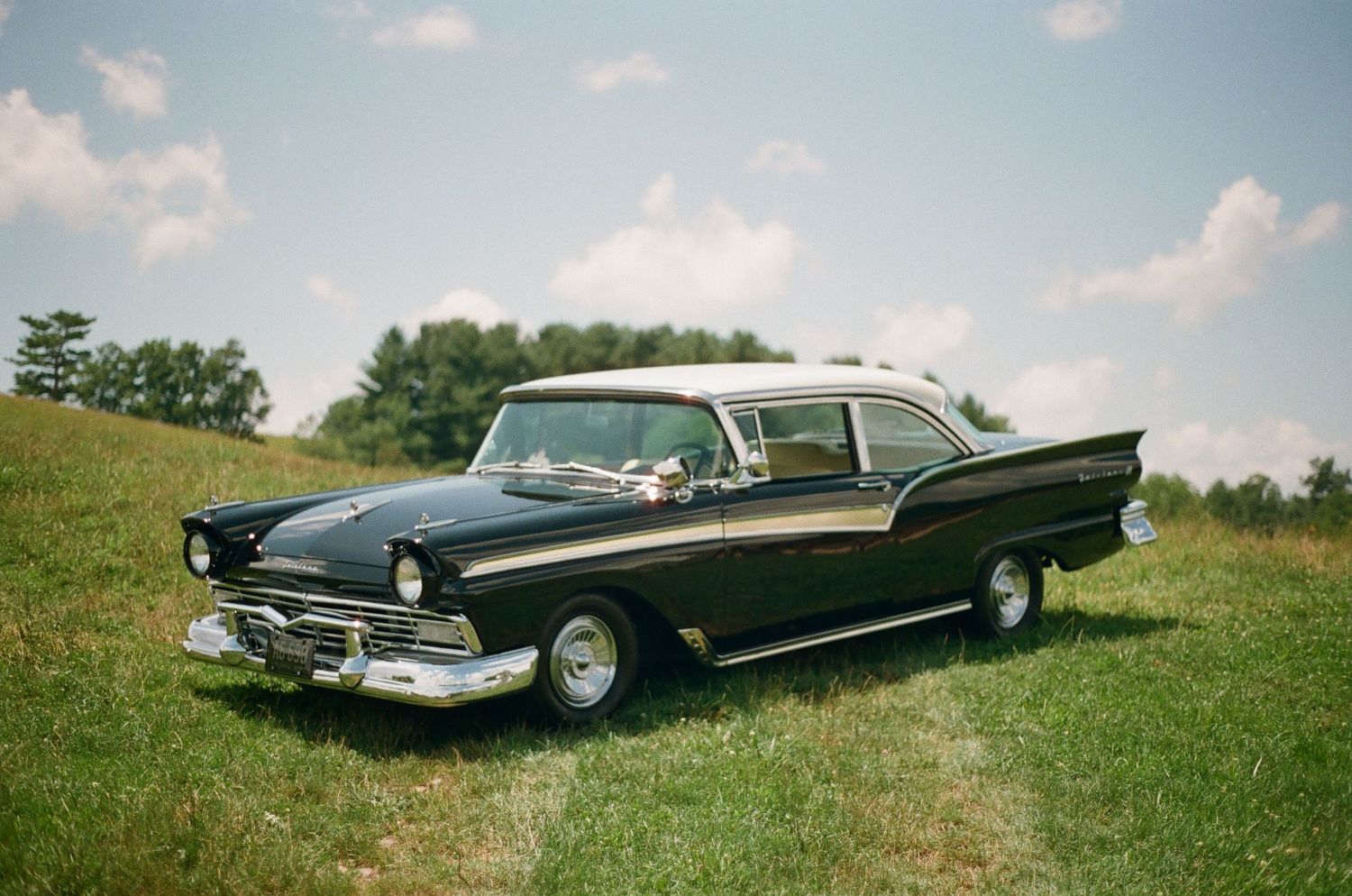 A sleek black 1950s Ford Fairlane with distinctive chrome trim sits in a sunny meadow.