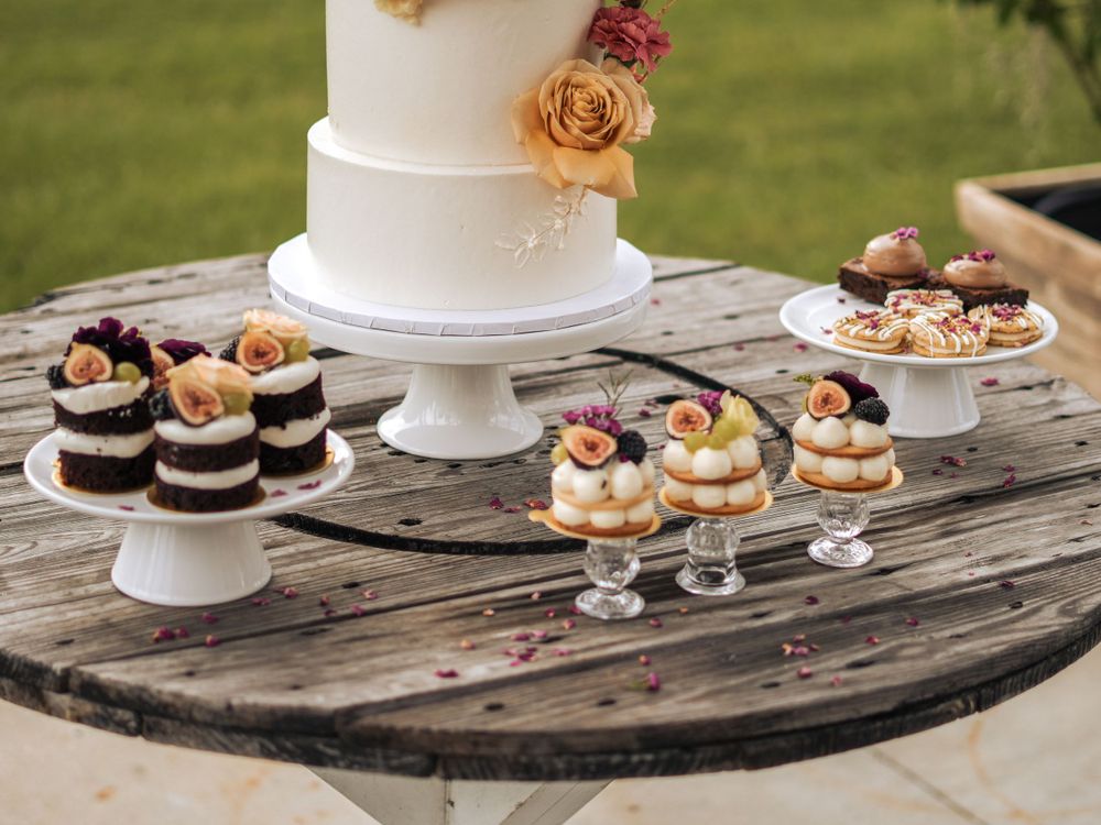 White tiered wedding cake with fresh flowers surrounded by delicate desserts on rustic wooden display.