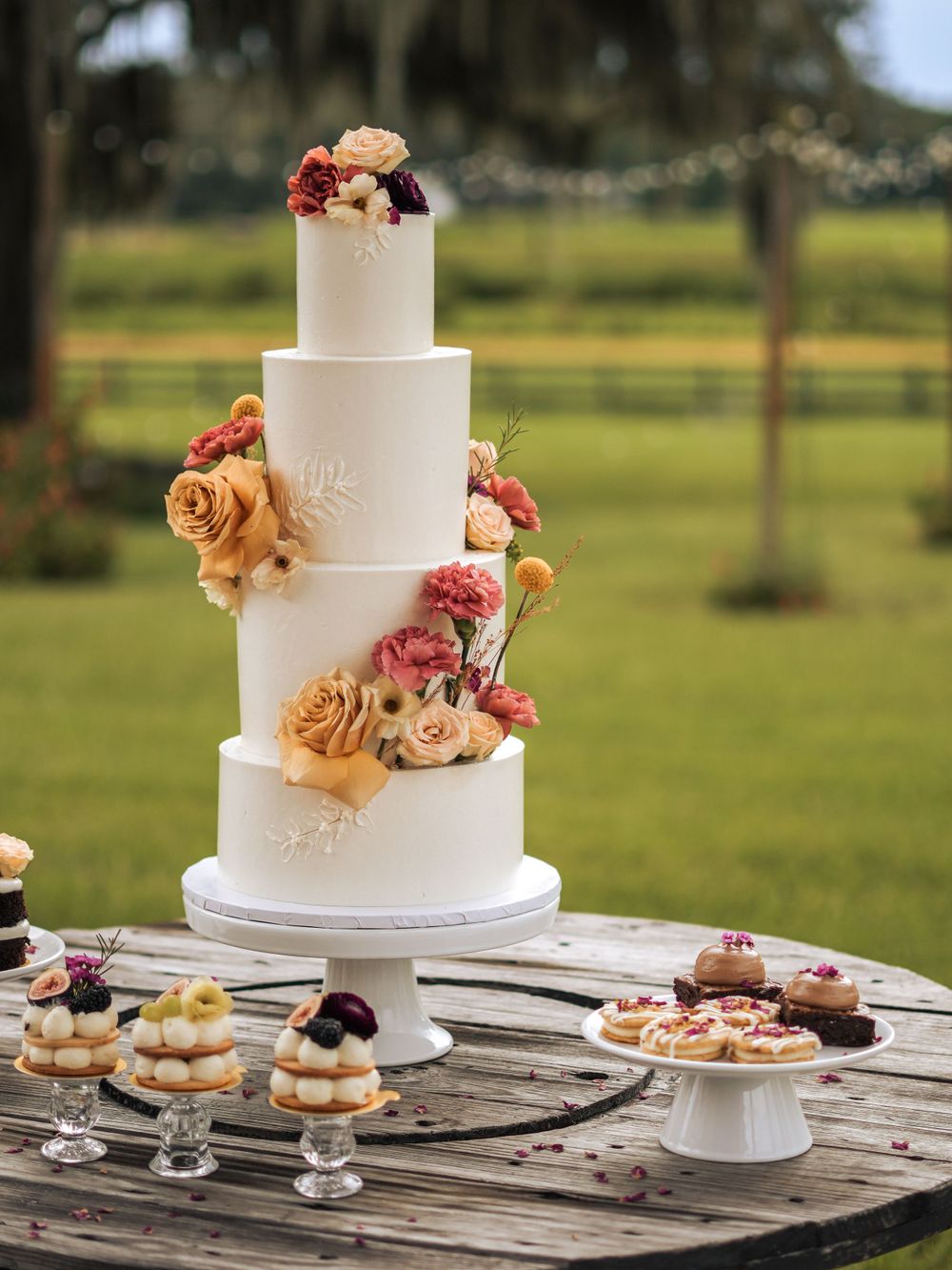 Elegant four-tier white wedding cake adorned with peach and pink roses on rustic dessert table.