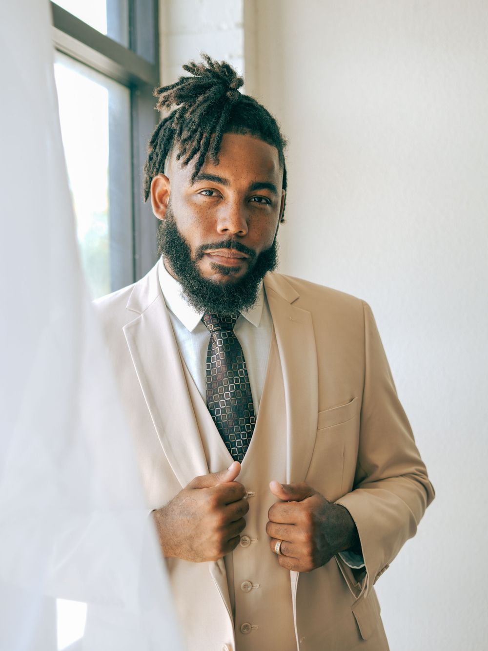 Professional wearing a beige suit and patterned tie stands confidently by a window in a modern setting.