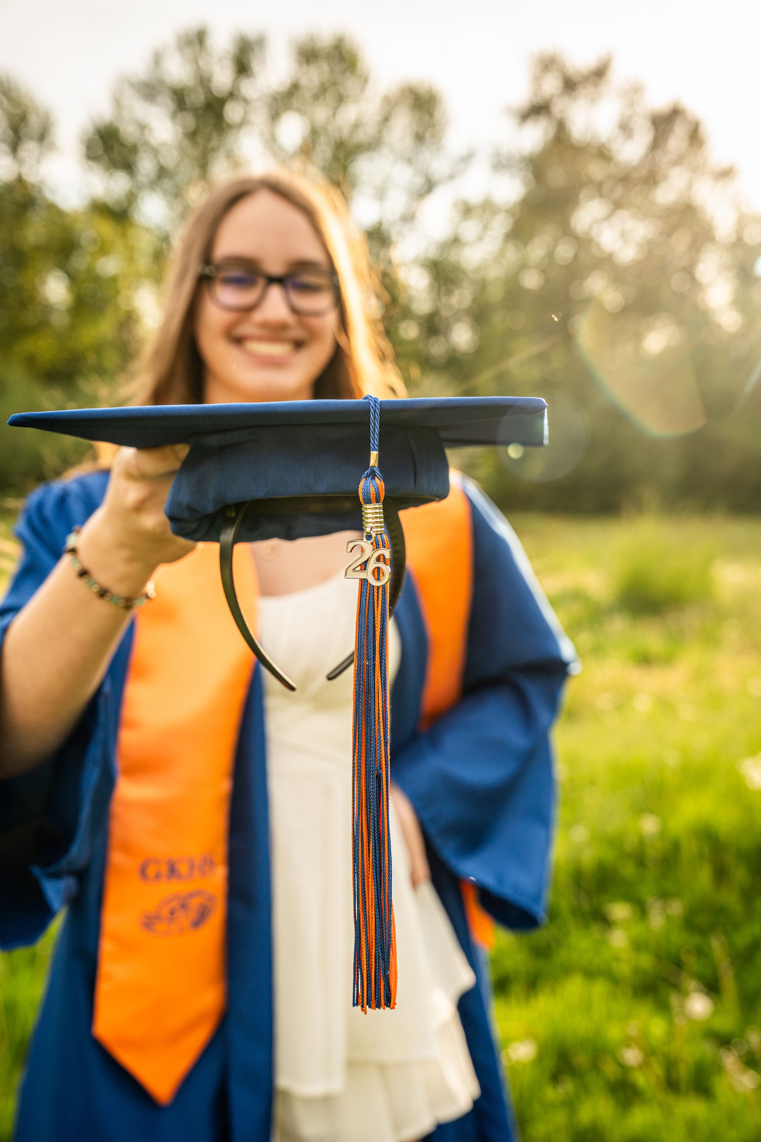 Smiling graduate in blue gown and orange stole holds cap toward camera outdoors in sunlit green field.