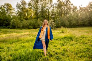 Smiling graduate in blue and gold cap and gown standing in a sunny green meadow with trees in background.