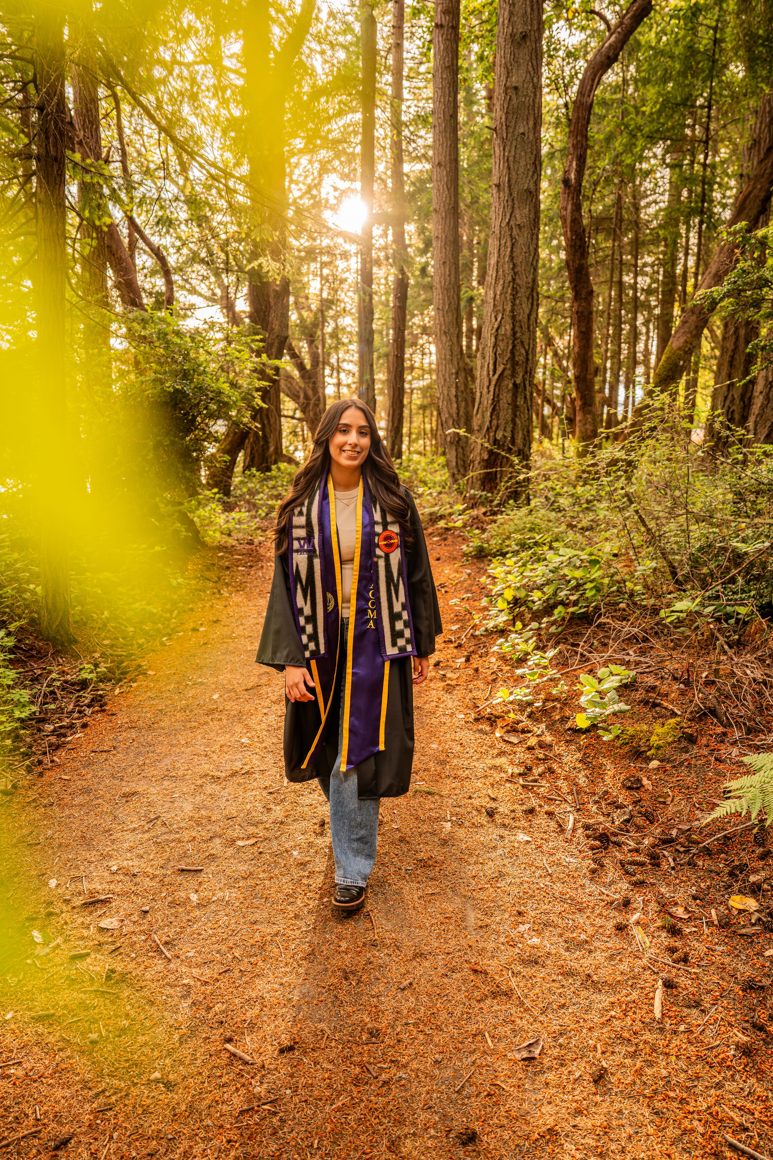 Graduate in cap and gown walks a sunlit forest path, wearing a colorful sash among towering redwood trees.