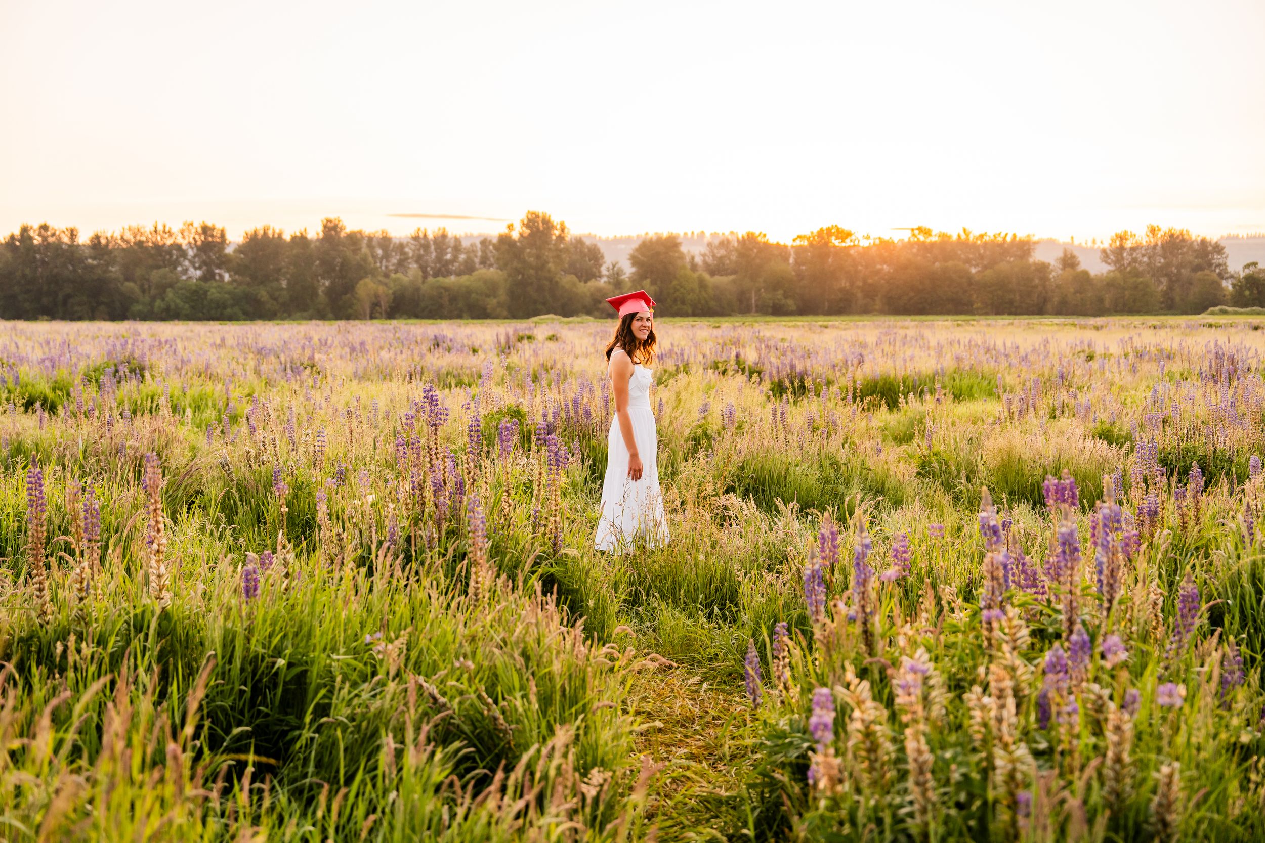 Woman in white dress standing in a lavender field at golden sunset with trees in the background.
