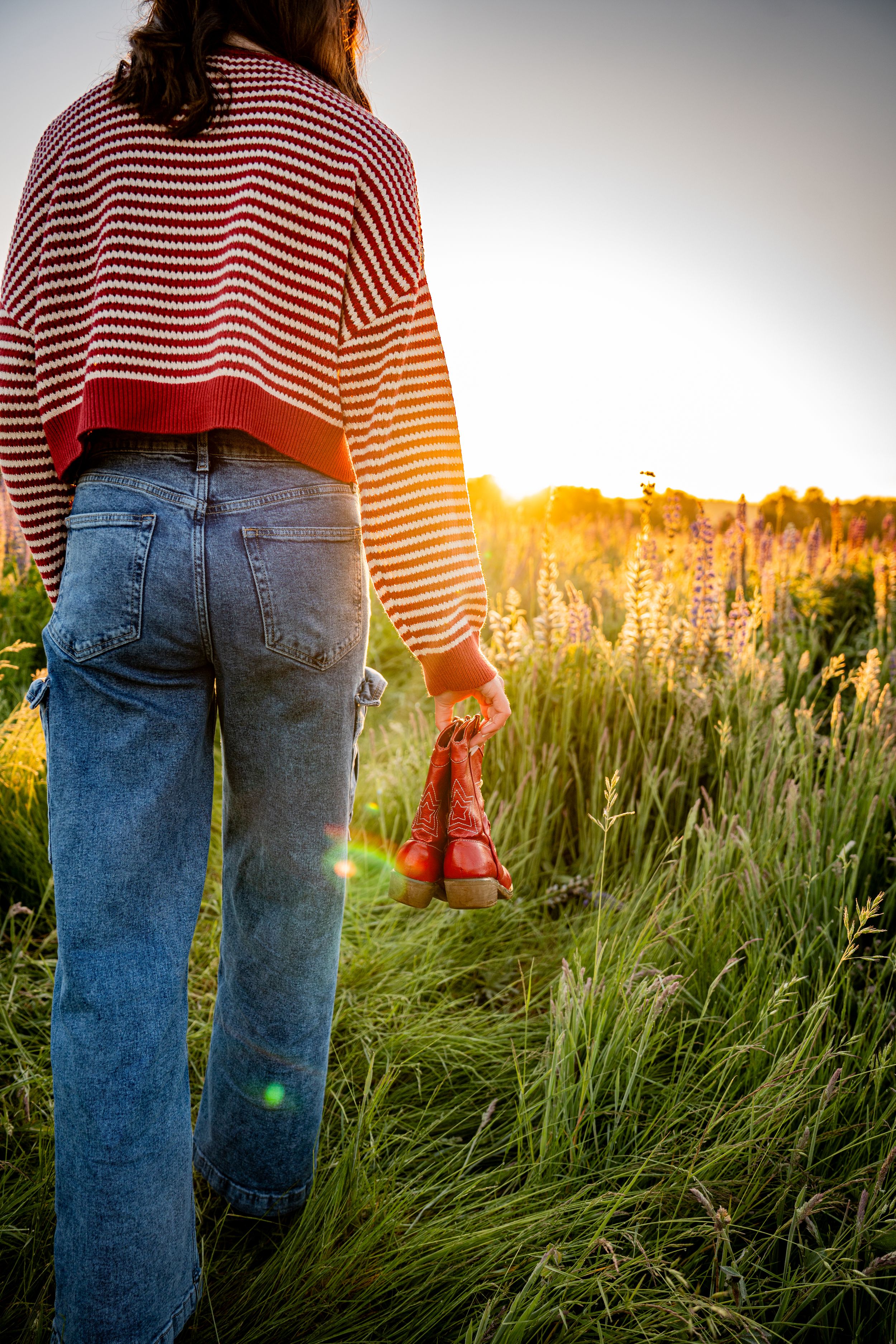 Woman in striped sweater and jeans holding red boots in a sunlit wildflower field at golden hour.