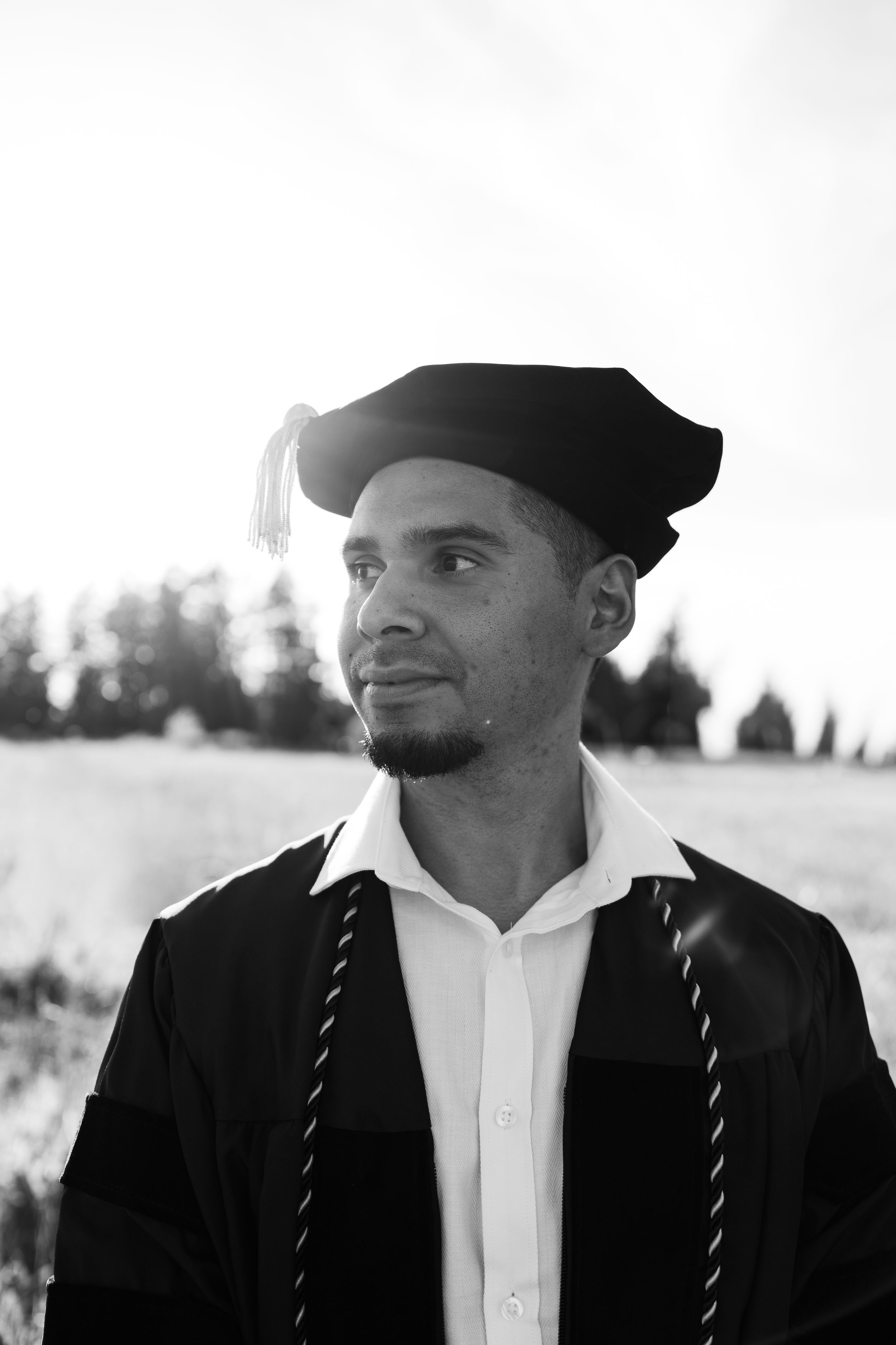 Black and white portrait of a young man in graduation cap and gown, gazing thoughtfully into the distance outdoors.
