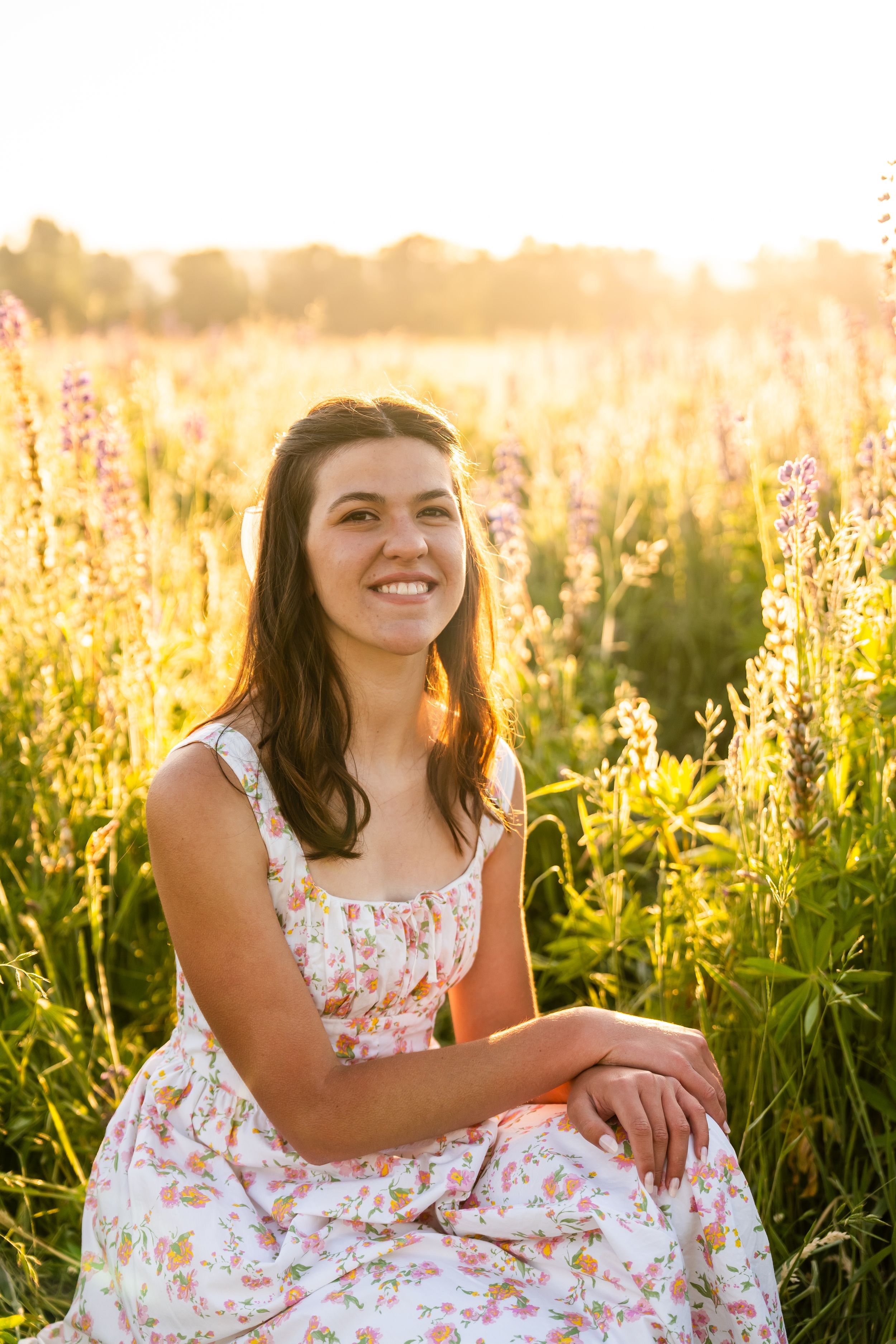 Smiling young woman in floral dress sitting in a sunlit wildflower field at golden hour.
