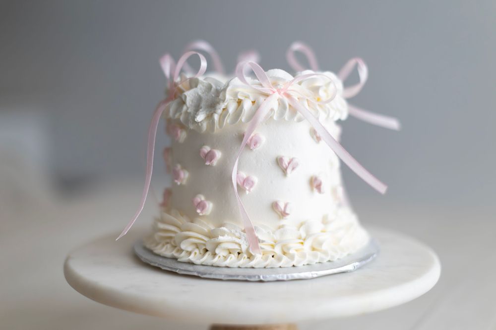Small white fondant cake decorated with delicate pink bows and ribbons sits on a white pedestal stand.