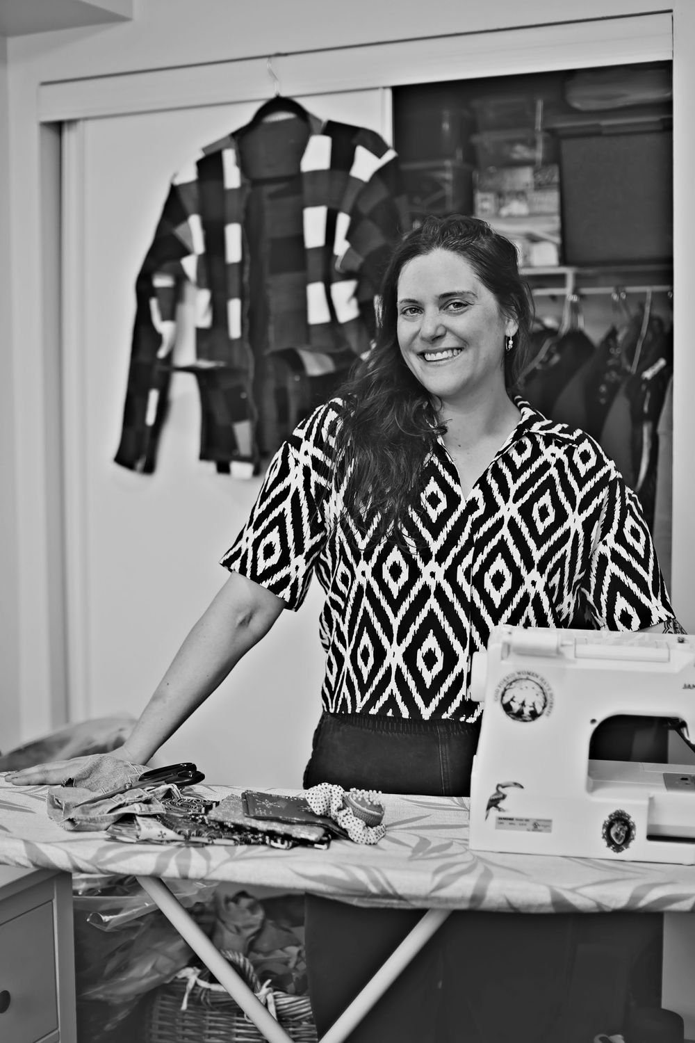 Smiling woman in patterned blouse stands at ironing board with sewing machine in black and white photo.