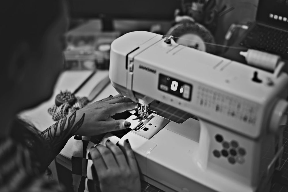 Person using a Brother sewing machine in a workshop, close-up black and white photo of hands guiding fabric.