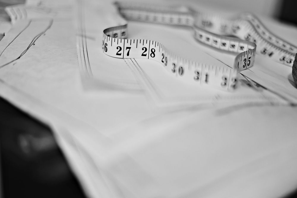 Black and white photo of a tailor measuring tape resting on white fabric pattern pieces on a work table.