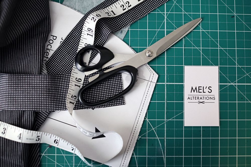 Mel's Alterations branding card with scissors, measuring tape, and fabric on a green cutting mat.