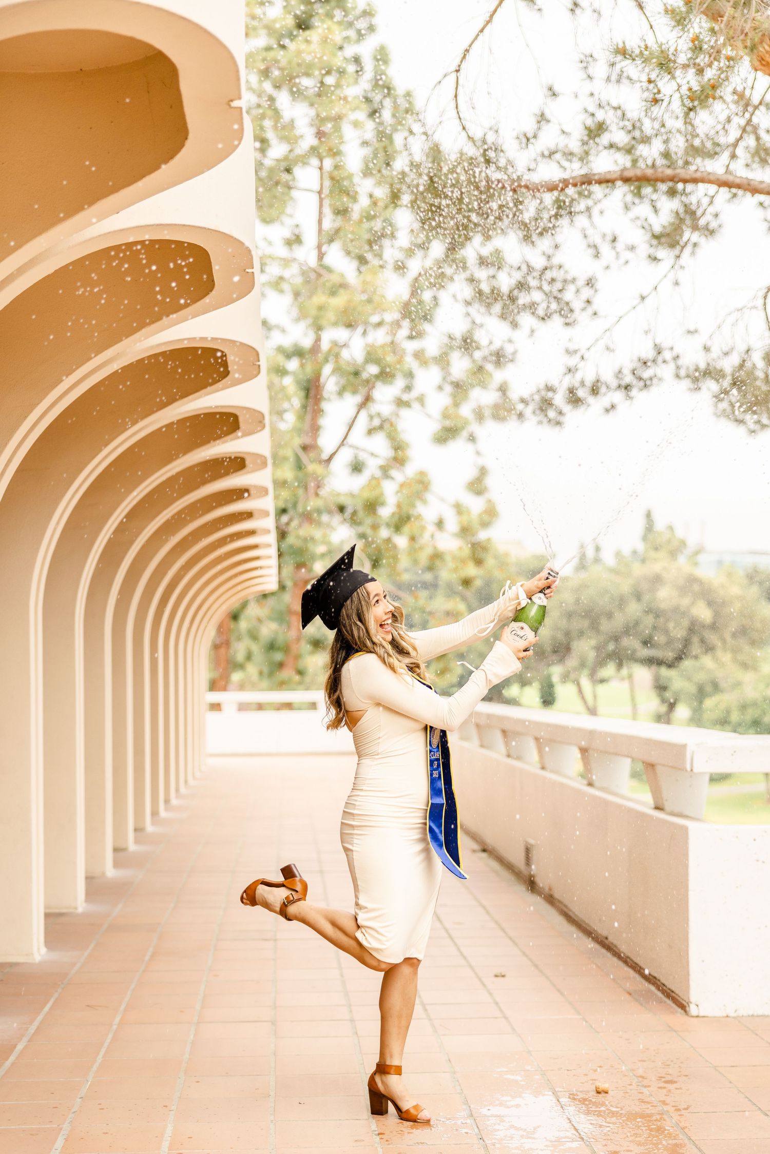 Graduate in blue dress poses playfully under arched colonnade walkway.