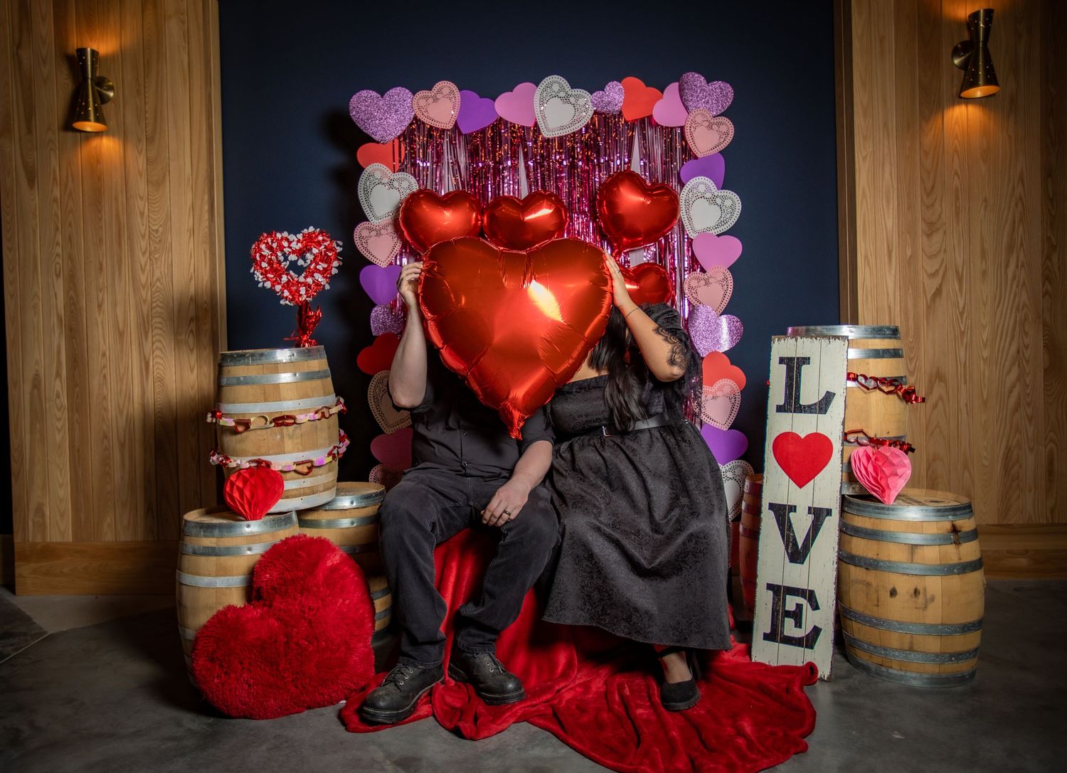 Valentine's Day themed wedding decor with red heart balloons, wooden barrels, and romantic lighting in a rustic venue.