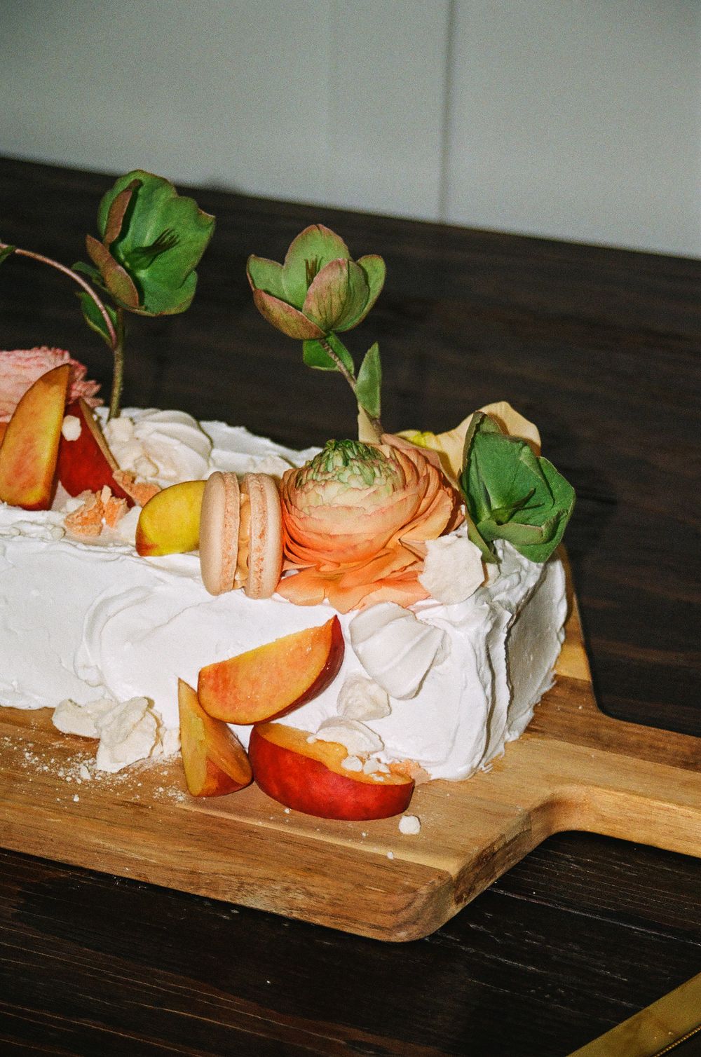 White frosted cake topped with peach slices, macarons, ranunculus flowers, and greenery on a wooden cutting board.