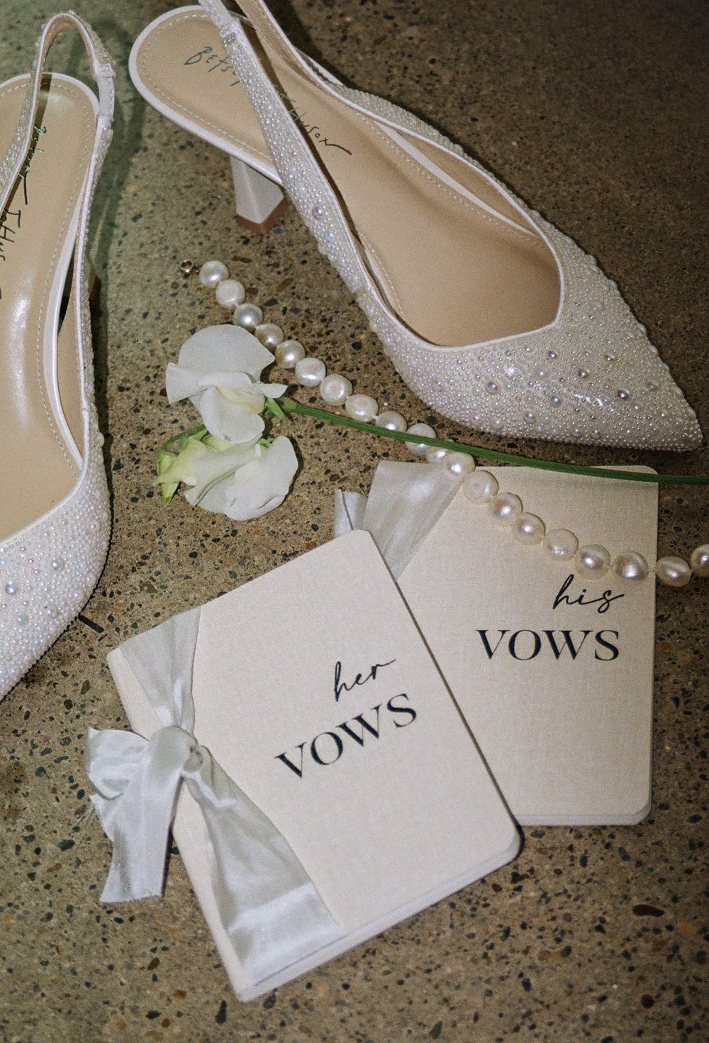 Wedding flat lay with his and her vow books, pearl necklace, white floral, and sparkly bridal slingback heels.