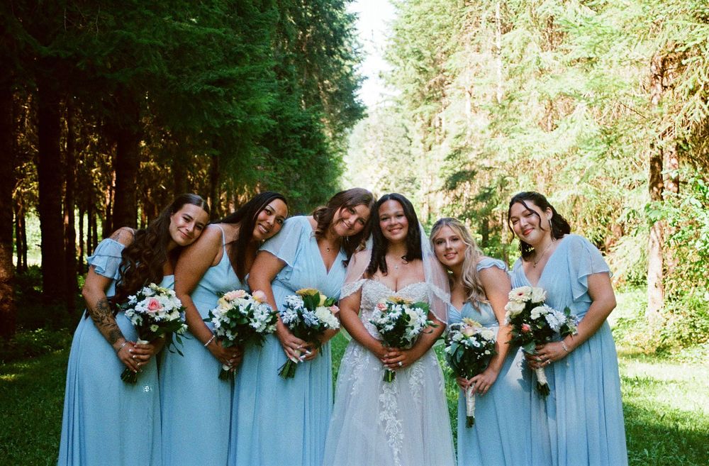 Bride in white gown poses with five bridesmaids in light blue dresses holding floral bouquets in a lush forest setting.