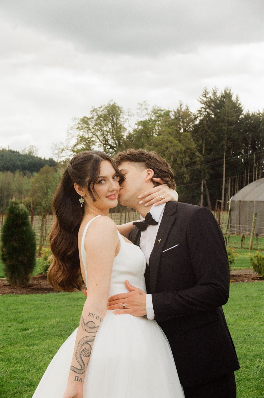 Bride in white gown smiles as groom in black tuxedo kisses her cheek at outdoor wedding venue.