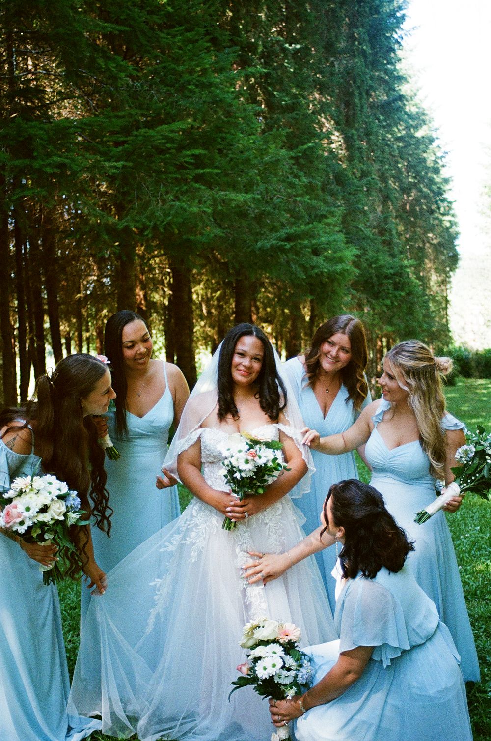 Bride in white lace gown and veil holds bouquet surrounded by bridesmaids in light blue dresses outdoors.