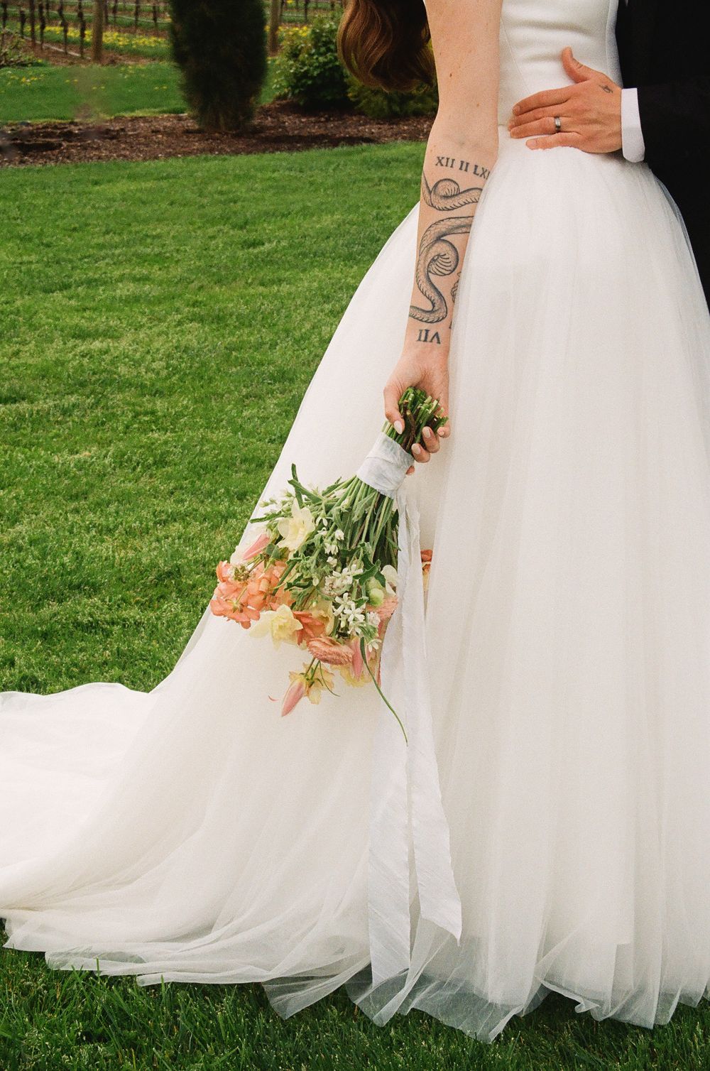 Bride in white tulle gown with floral tattoo holding peach bouquet, embraced by groom in suit on green lawn.