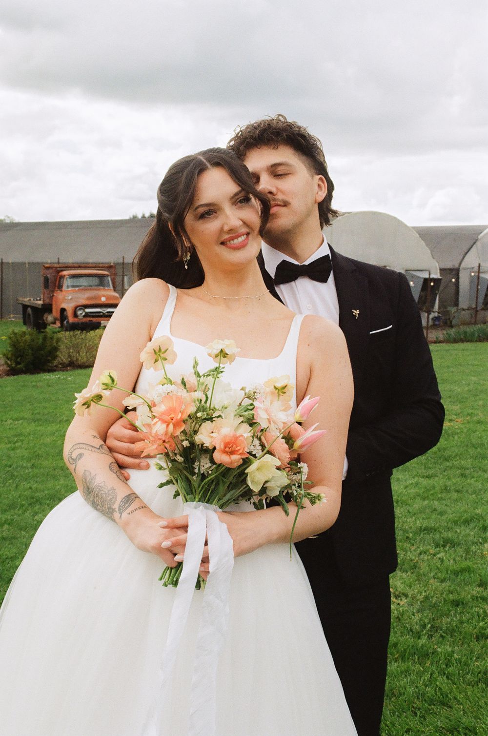 Bride and groom pose outdoors, she holds a peach floral bouquet in a white gown as he kisses her cheek in a black tux.