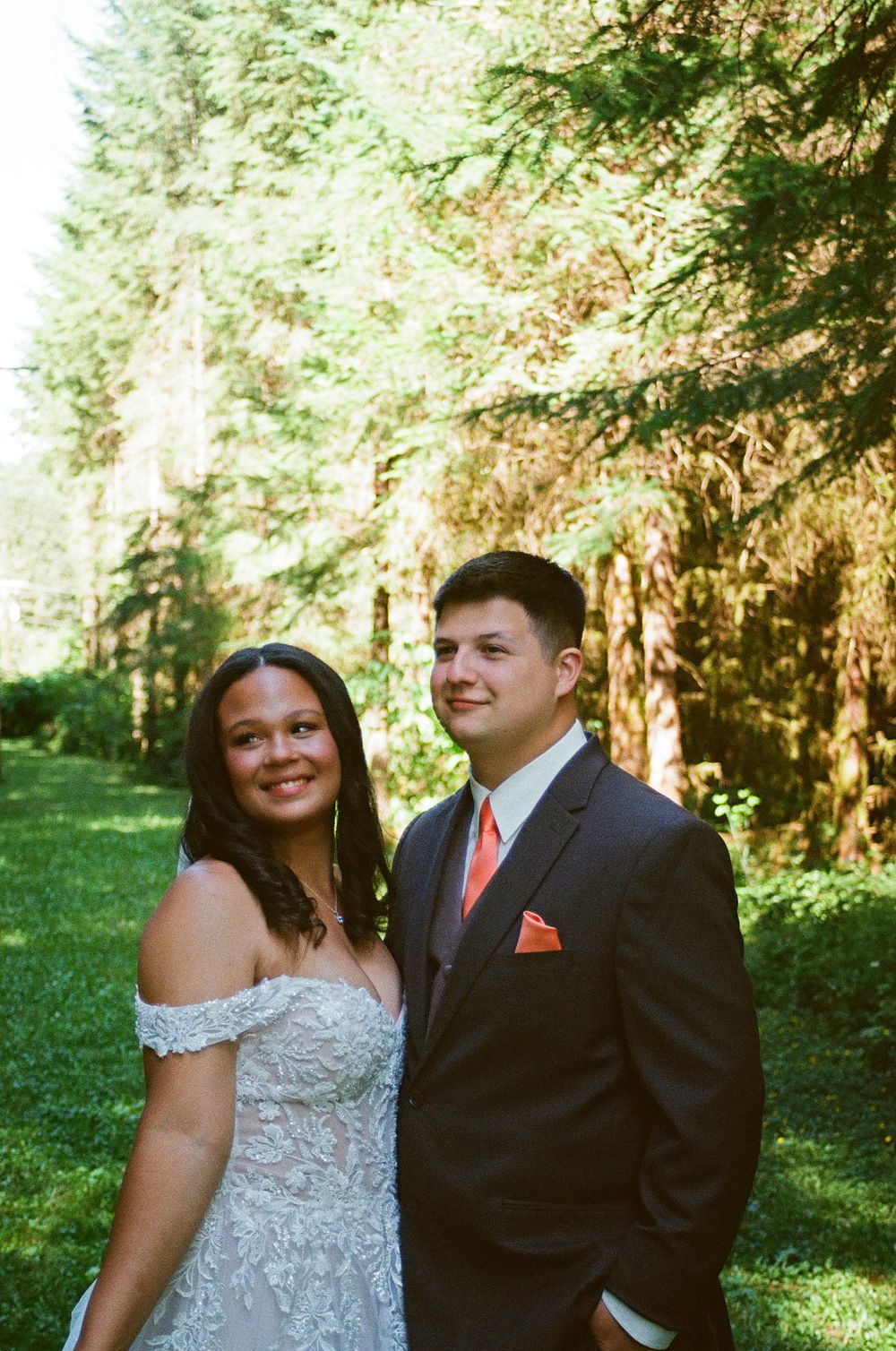 Bride and groom smiling together in a lush green forest setting, bride in lace off-shoulder gown, groom in navy suit.