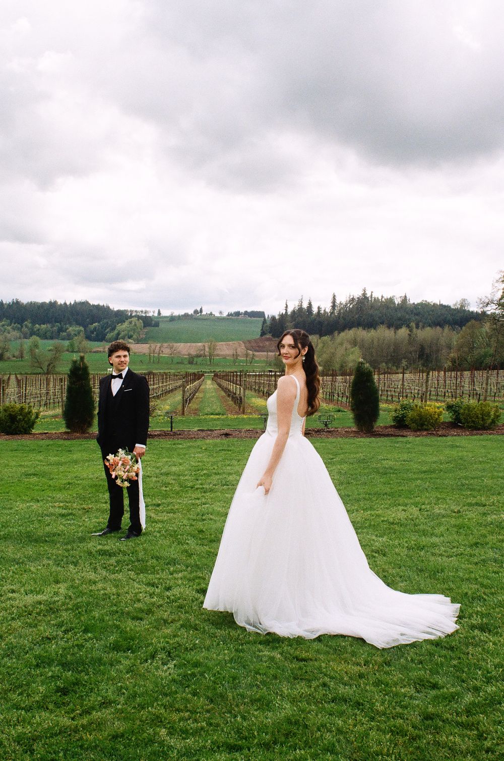 Bride in white ballgown and groom in tuxedo pose on green lawn at vineyard wedding venue under cloudy sky.