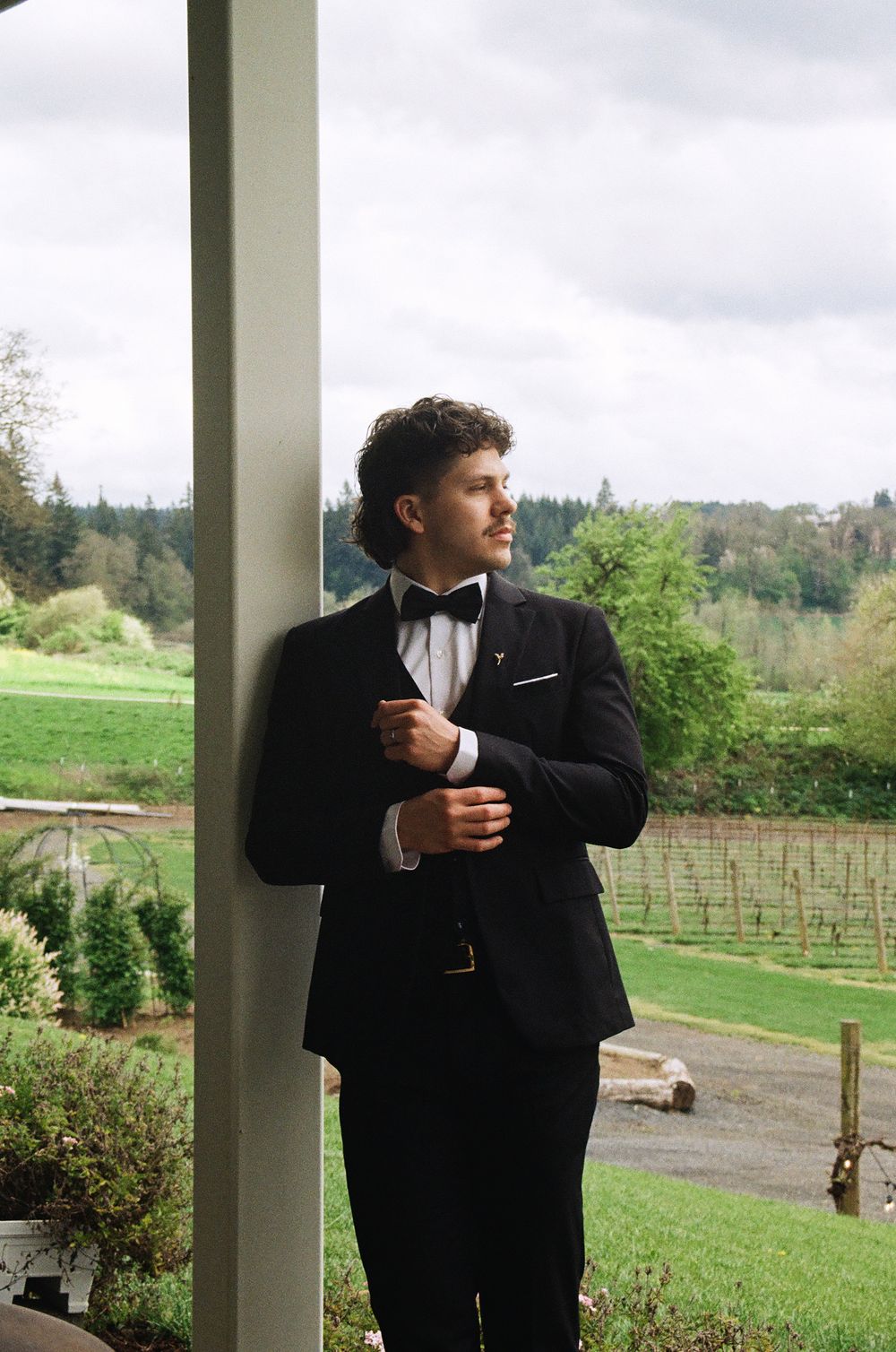 Groom in black tuxedo and bow tie adjusting cufflinks by a pillar overlooking a lush green vineyard.