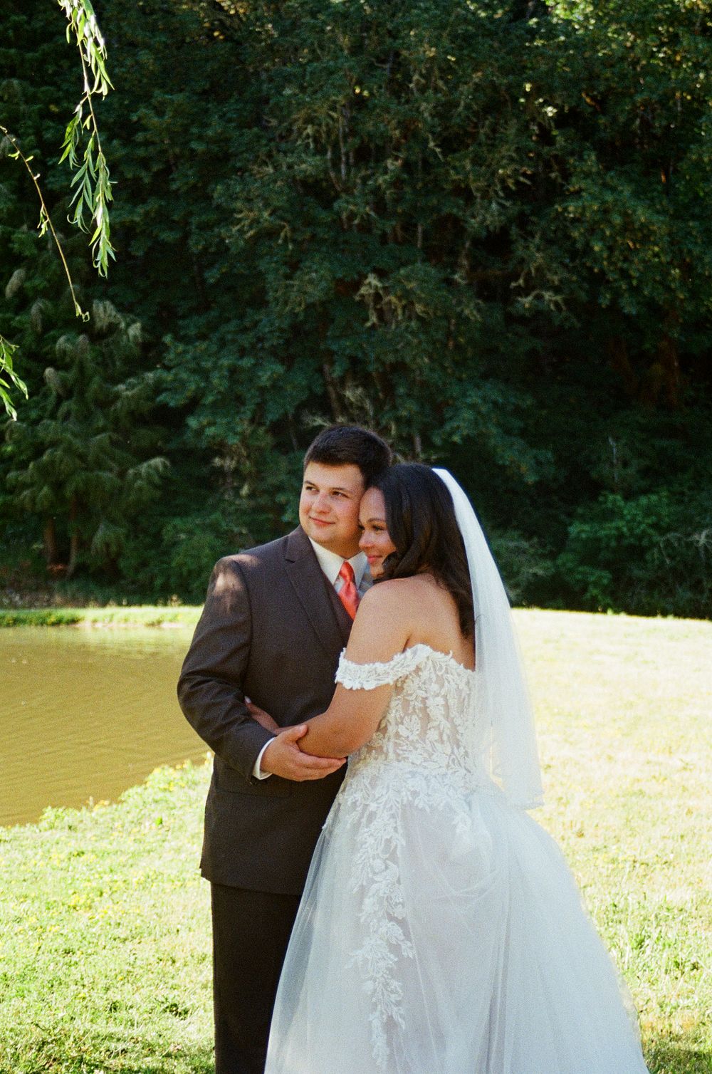 Bride in lace off-shoulder wedding gown with veil embraces groom in dark suit by a serene pond outdoors.