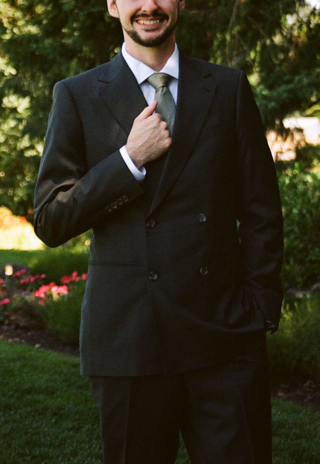 Smiling man in dark double-breasted suit and green tie posing outdoors in a garden setting.