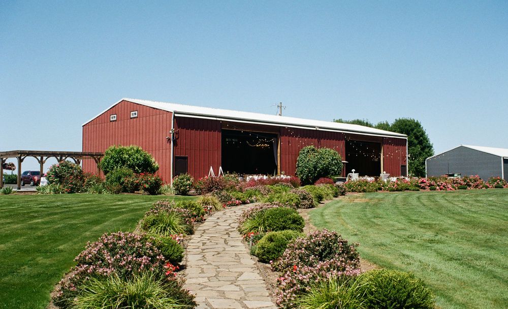 Red barn with open doors, surrounded by lush gardens and a stone path on a sunny day.