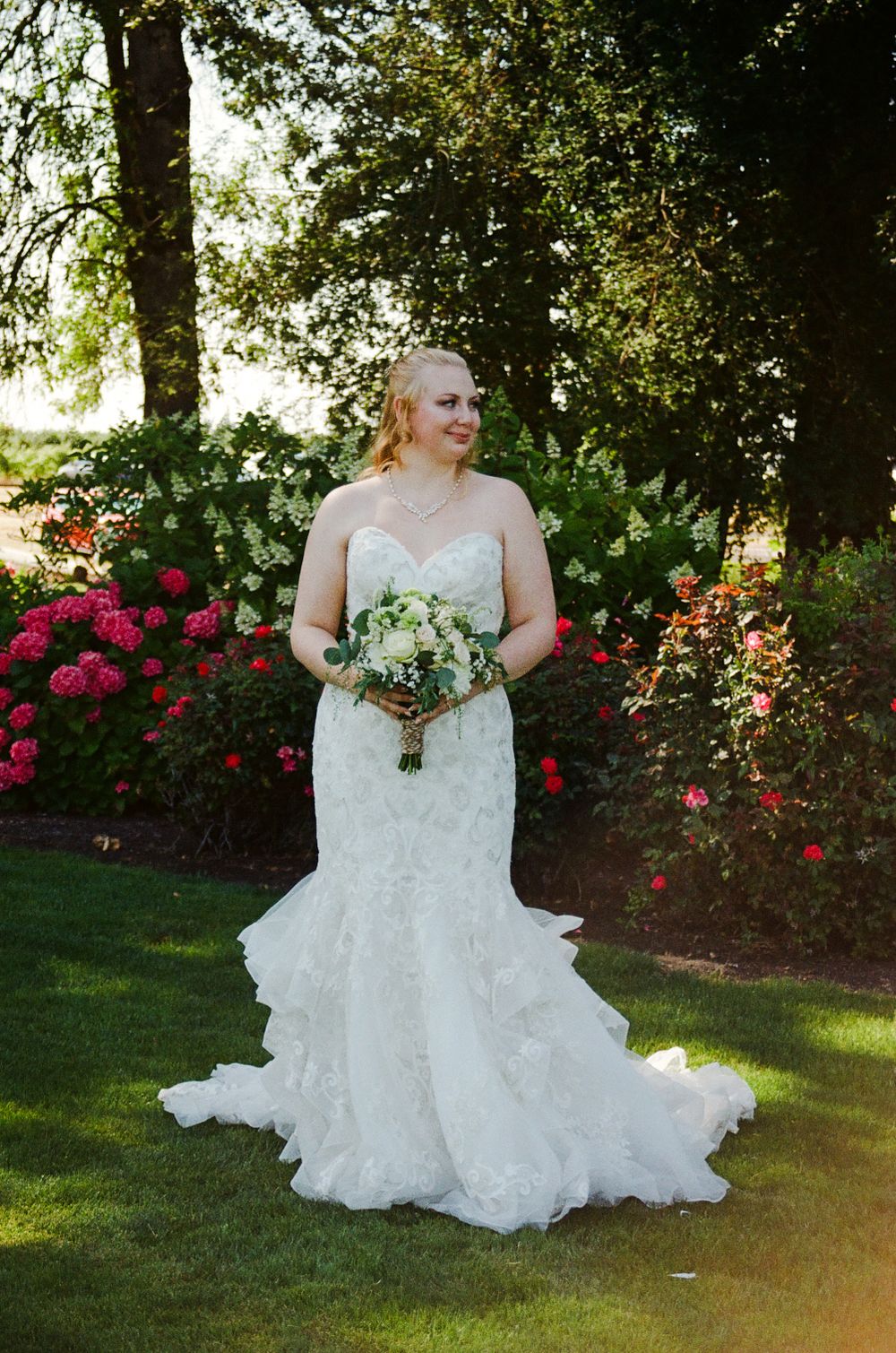 Bride in a lace mermaid wedding gown holding a white bouquet, standing in a garden with colorful roses.