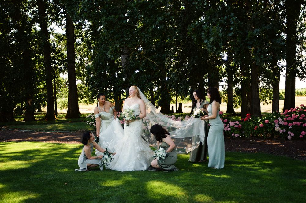 Bride in white gown surrounded by bridesmaids in sage green dresses on lush lawn with tall trees and pink flowers.