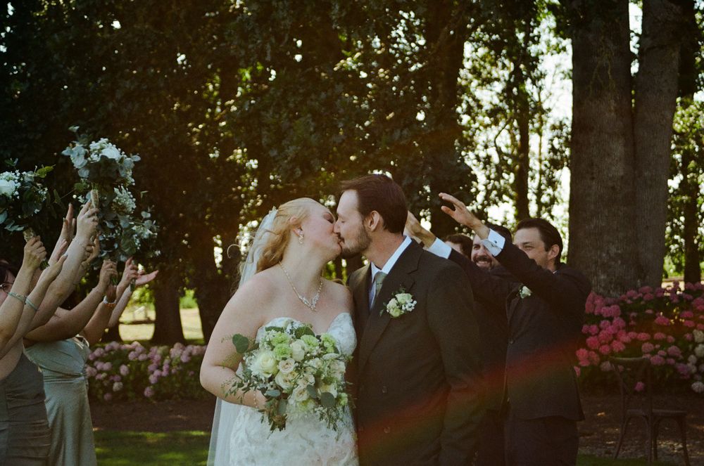 Bride and groom share a first kiss outdoors surrounded by wedding party with lush green trees in background.