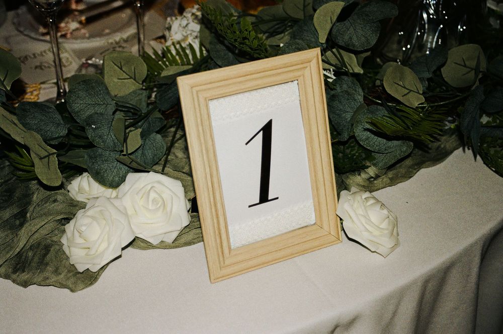 Wooden framed table number one displayed on white tablecloth with white roses and green eucalyptus garland centerpiece.