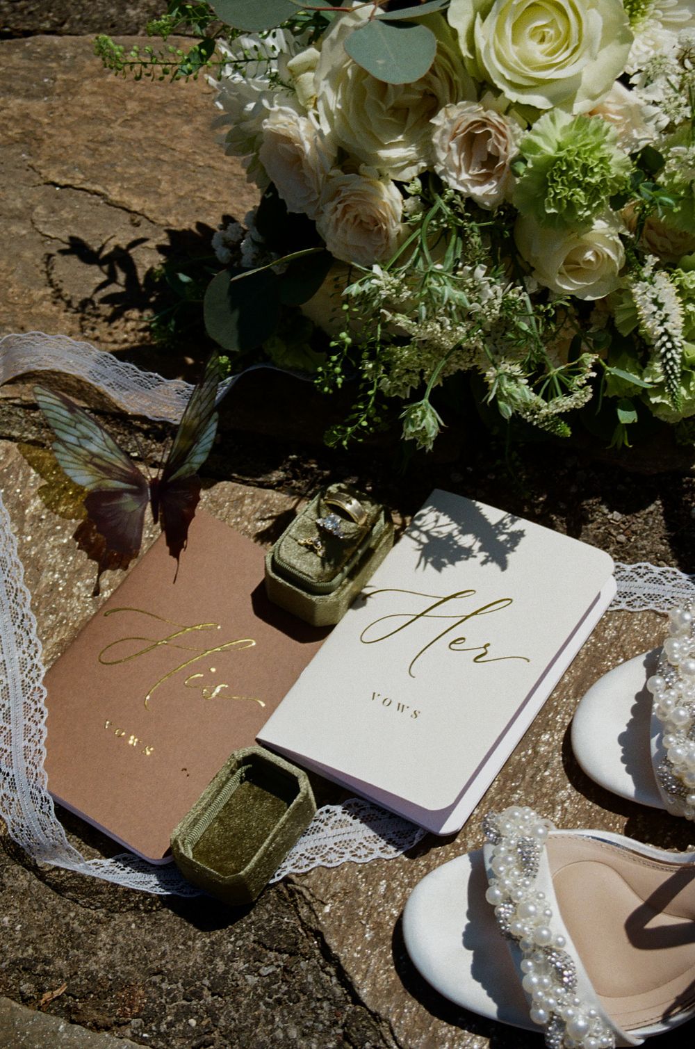 Wedding flat lay with vow books labeled His and Hers, ring boxes, lace ribbon, pearl heels, and white floral bouquet on stone.