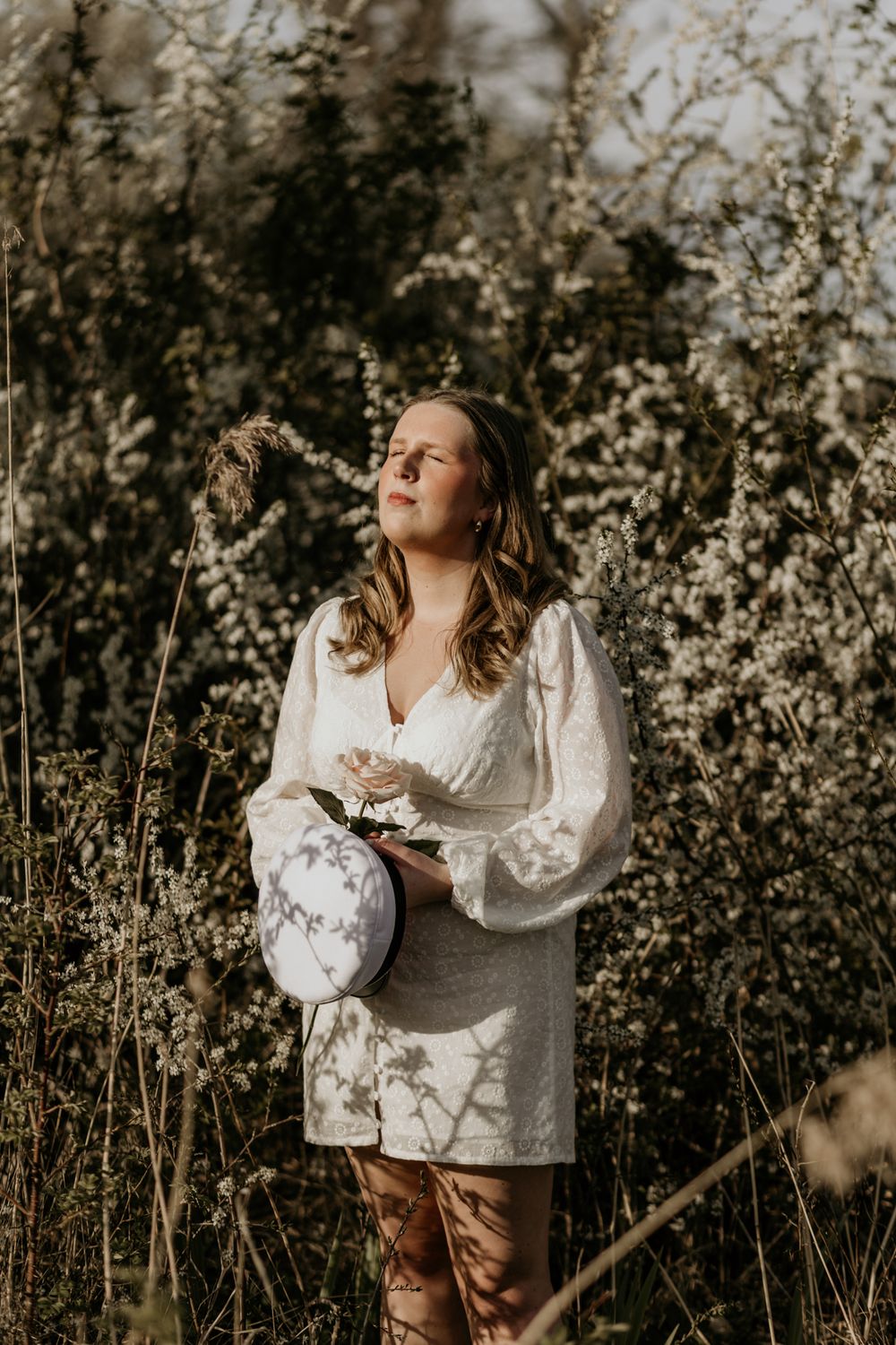 A person in a white dress poses with a decorative drum in a sunlit field of wildflowers.
