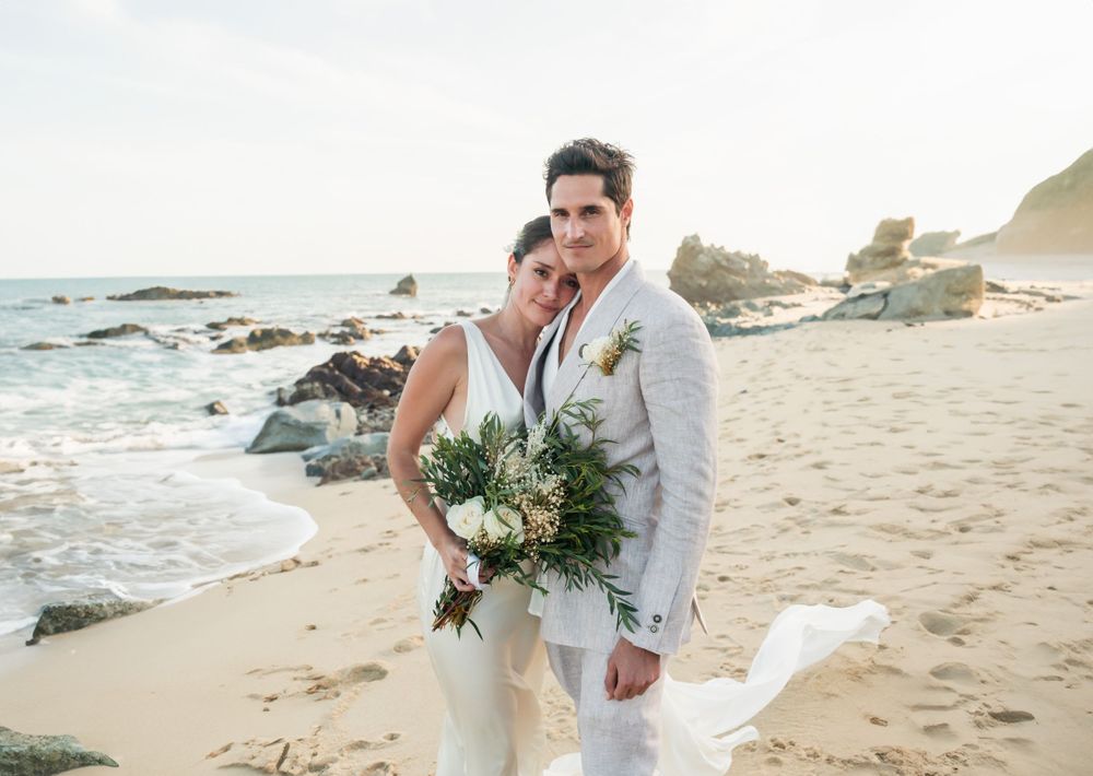 A romantic beach wedding portrait with rocky coastline and crashing waves at sunset.