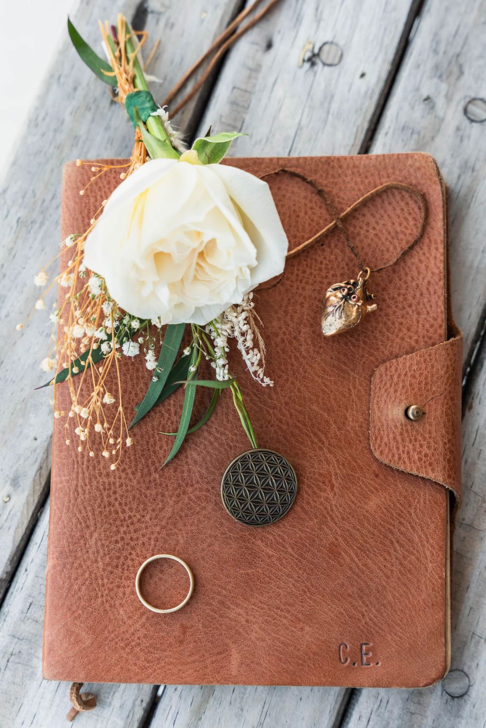 A white rose and baby's breath rest on a brown leather journal with initials and a wedding ring.