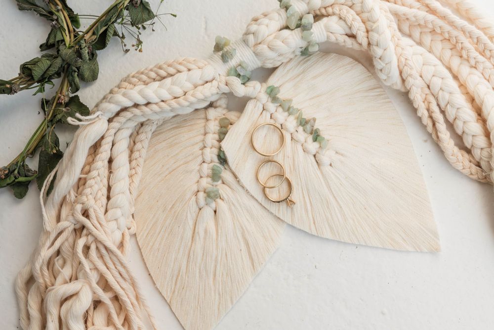 Wedding rings styled with macrame leaves and dried botanicals on cream background.